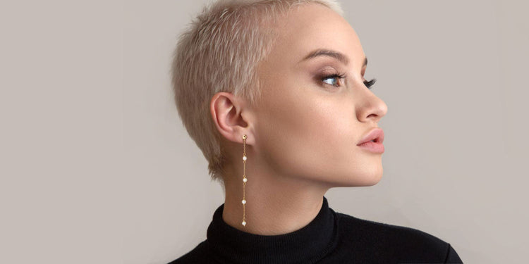 Frau mit kurzen platinblonden Haaren trägt extravagante lange goldene Ohrhänger mit mehreren funkelnden Steinchen, im Profil vor neutralem Hintergrund in schwarzem Oberteil