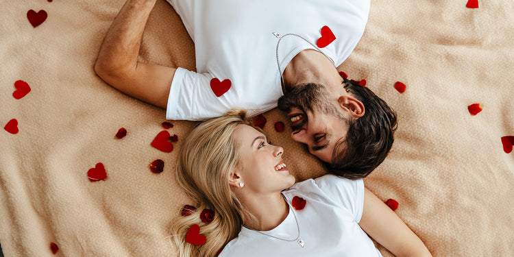 Glückliches Paar liegt auf einer beigen Decke, umgeben von roten Rosenblättern und Herzen. Die blonde Frau und der dunkelhaarige Mann tragen weiße Kleidung und lächeln sich verliebt an. Beide tragen zarte Halsketten mit einem geteilten Herz-Anhänger.