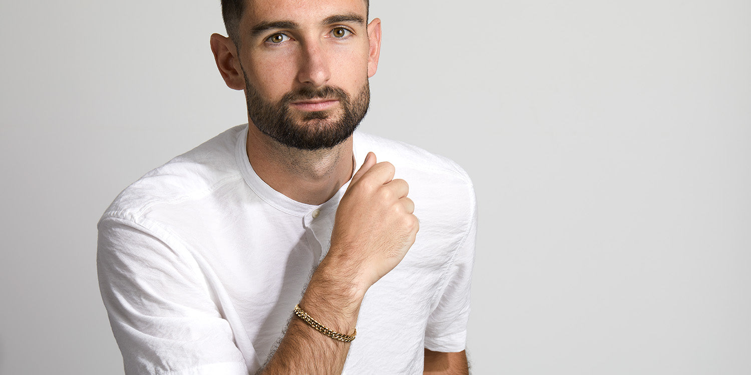 Mann mit kurzem Haar und Bart in weißem Langarmshirt trägt goldenes Panzerarmband 