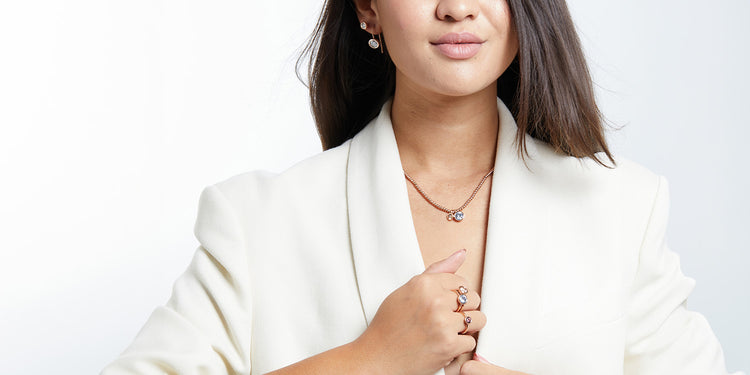 Elegante Frau mit braunen Haaren in weißem Blazer trägt goldene Halskette mit blauem Anhänger, goldene Ohrringe mit blauen Steinen und mehrere goldene Ringe 